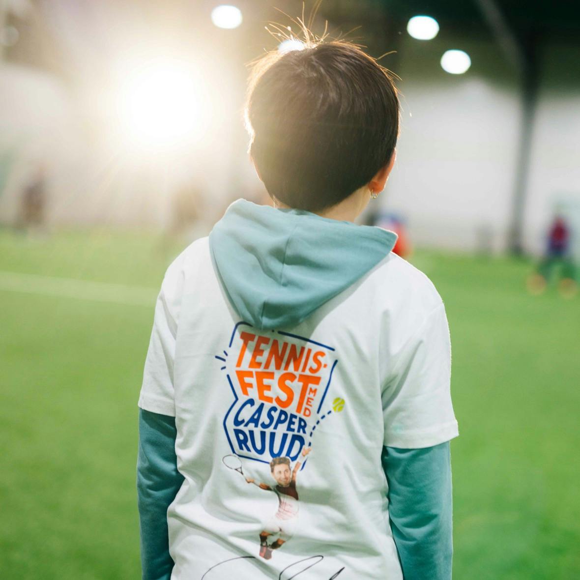 Child with short dark hair is standing with their back to the camera on an indoor fotball field. He is wearing a signed t-shirt by Casper Ruud 